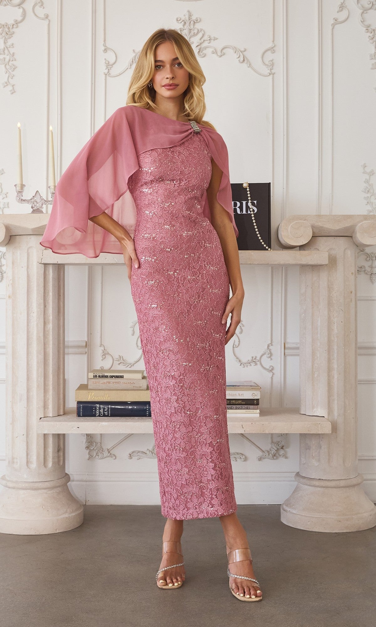 A woman stands indoors in a Dusty Rose Long Prom Dress: Amoris T906 with sequin-lace details and matching chiffon cape overlay, posing before a decorated white mantel lined with books.
